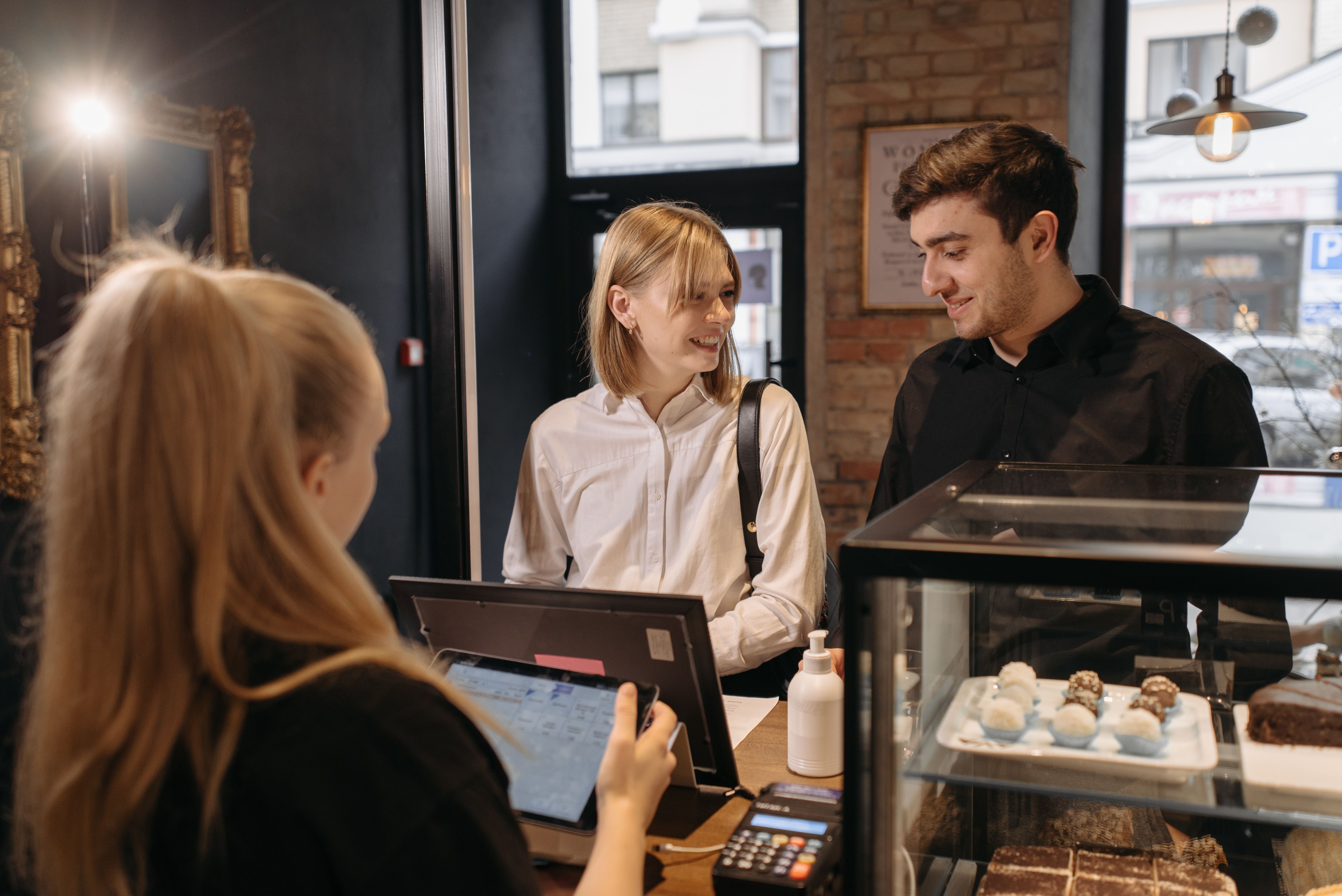 Two customers being helped at a small bakery counter while a staff member rings them up on a tablet.